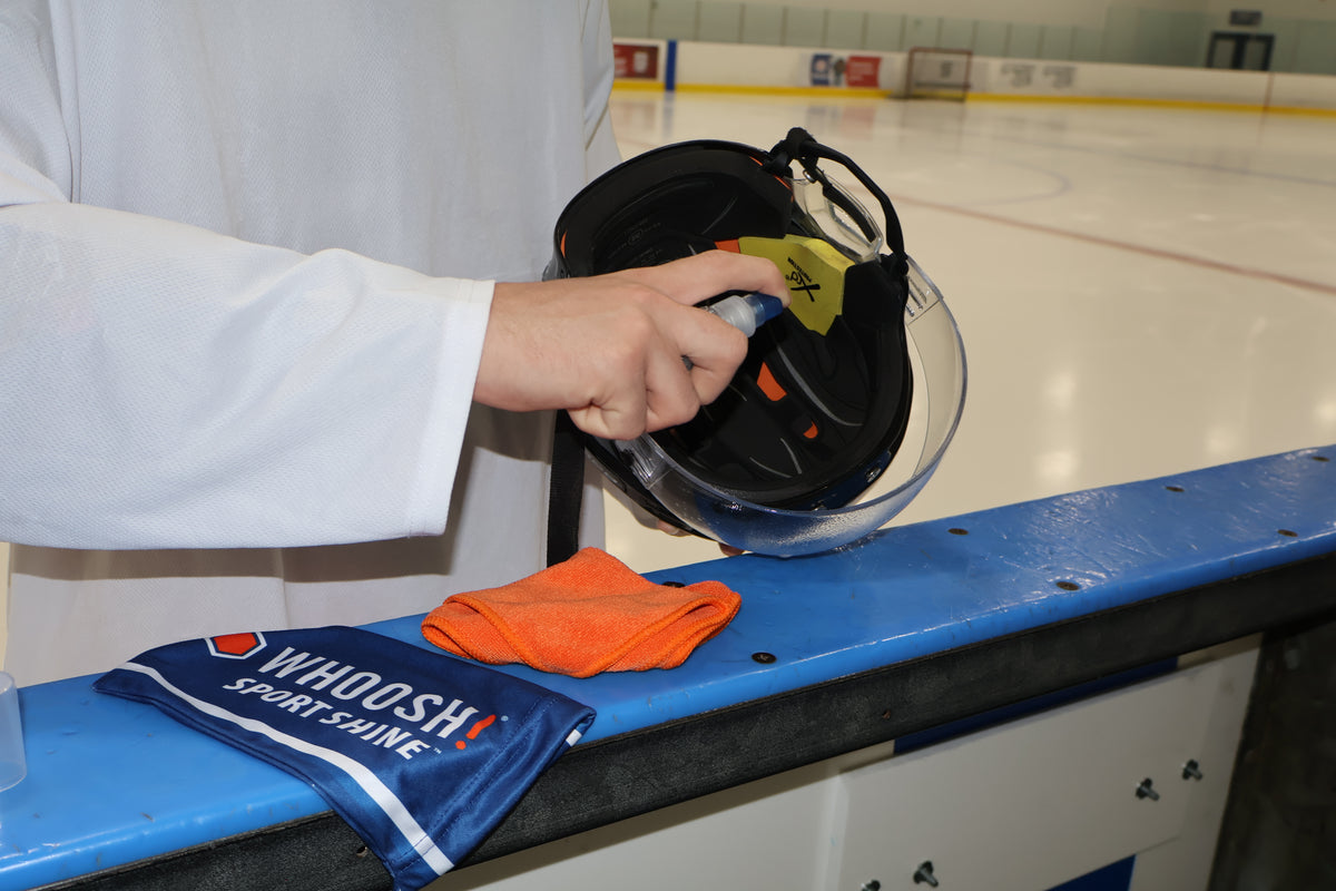 Person cleaning a hockey visor by spraying WHOOSH! Sport Shine directly on the visor. The cloth and carry bag are sitting next to the helmet.