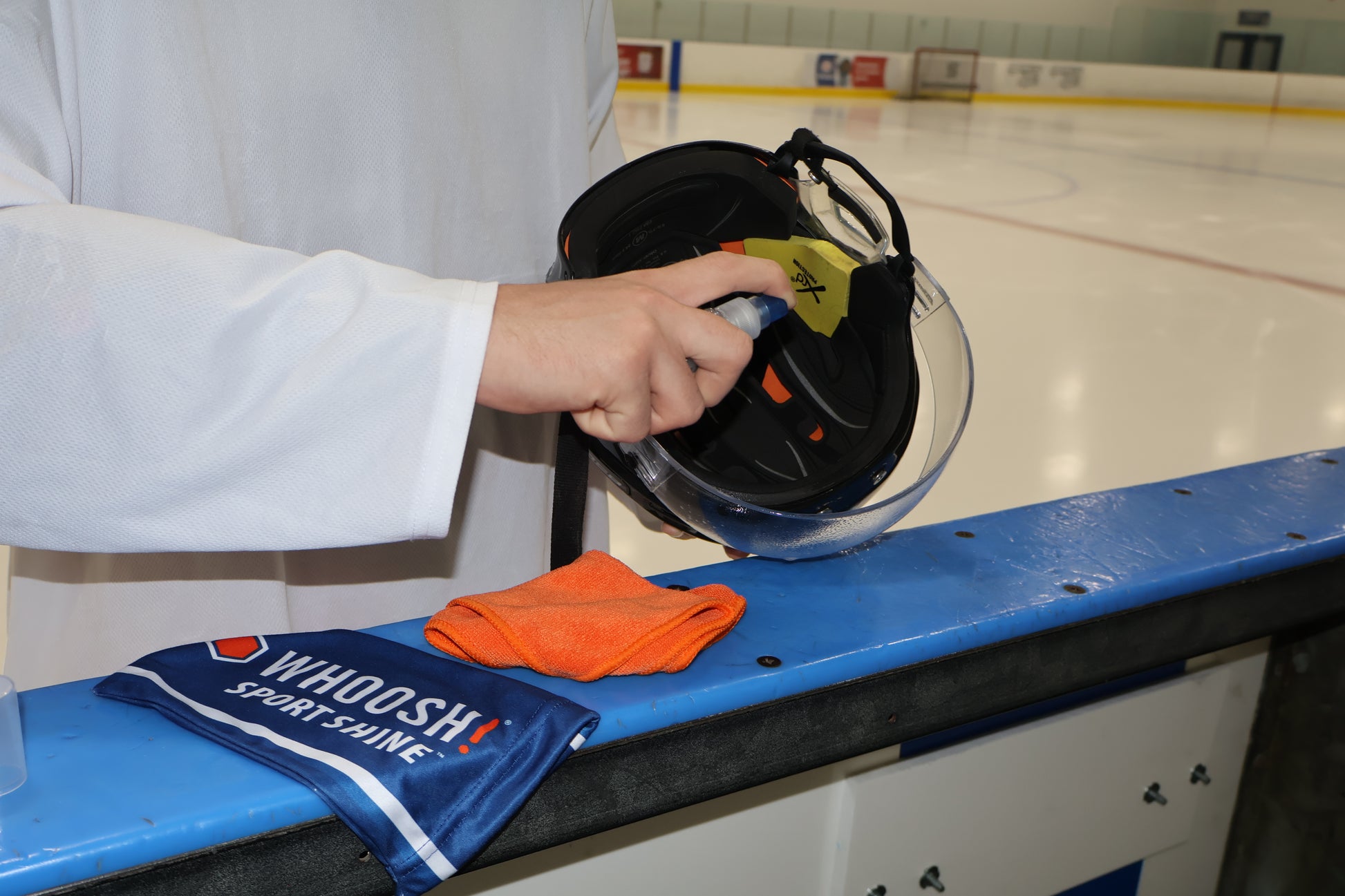 Person cleaning a hockey visor by spraying WHOOSH! Sport Shine directly on the visor. The cloth and carry bag are sitting next to the helmet. 