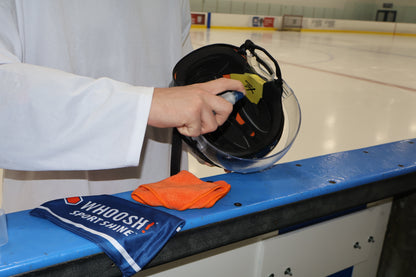 Person cleaning a hockey visor by spraying WHOOSH! Sport Shine directly on the visor. The cloth and carry bag are sitting next to the helmet. 