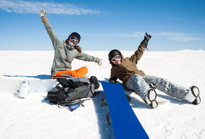 Two people with snowboards in a snowy landscape with a clear blue sky. One person is holding a WHOOSH! sport bottle and the other has the carry case in front of them.