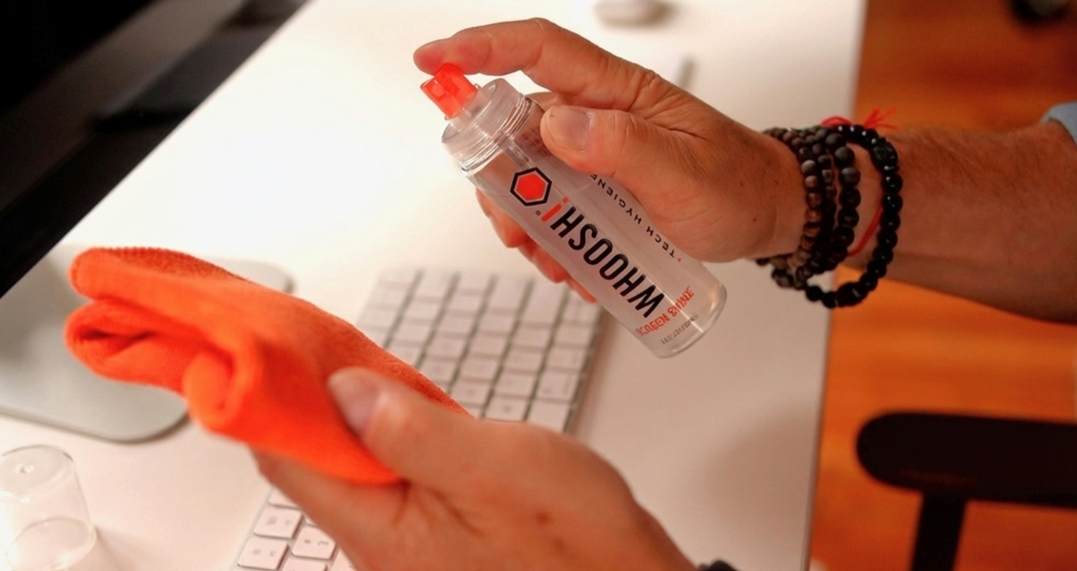 Person cleaning a keyboard with a spray bottle labeled 'WHOOSH!' on a desk.