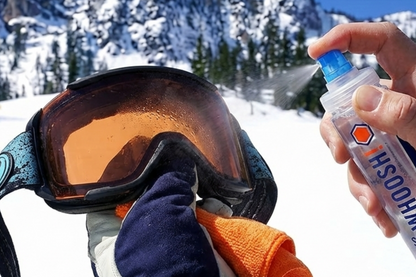 Person spraying WHOOSH! Sport Shine directly on ski goggles with cloth in hand, in a snowy mountain setting.