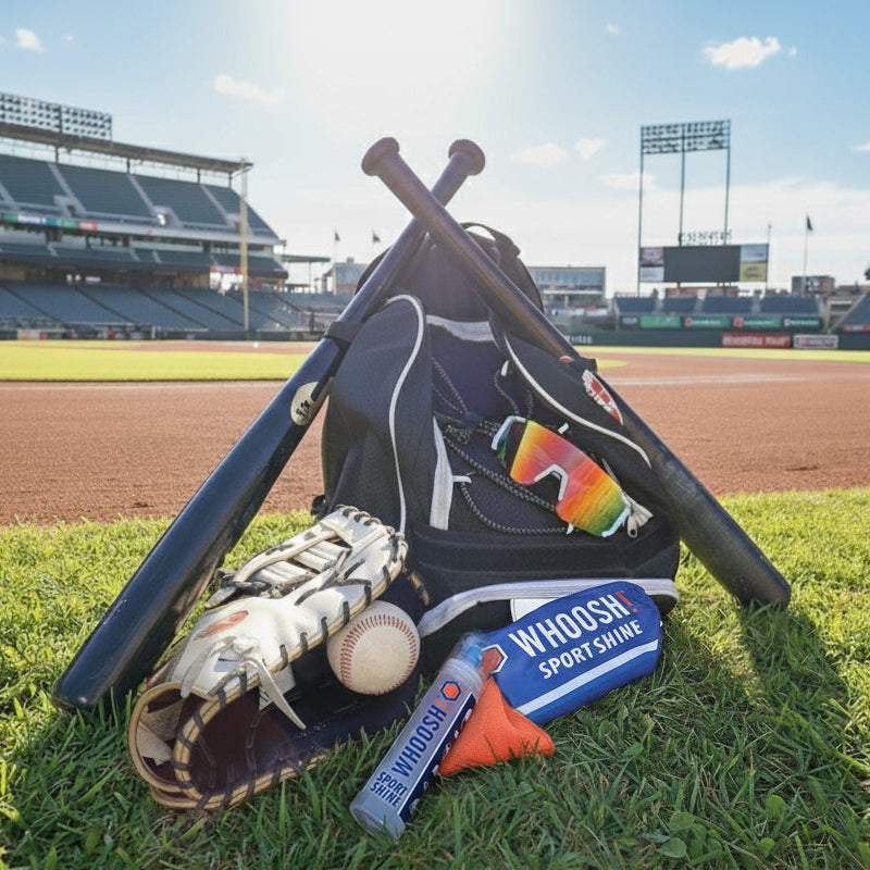 Baseball glove with ball in glove, bat, and sport glasses sitting on the baseball bag. WHOOSH! Sport Shine carry bag, spray and cloth are sitting infront of the bag in the grass, with a baseball stadium in the background.